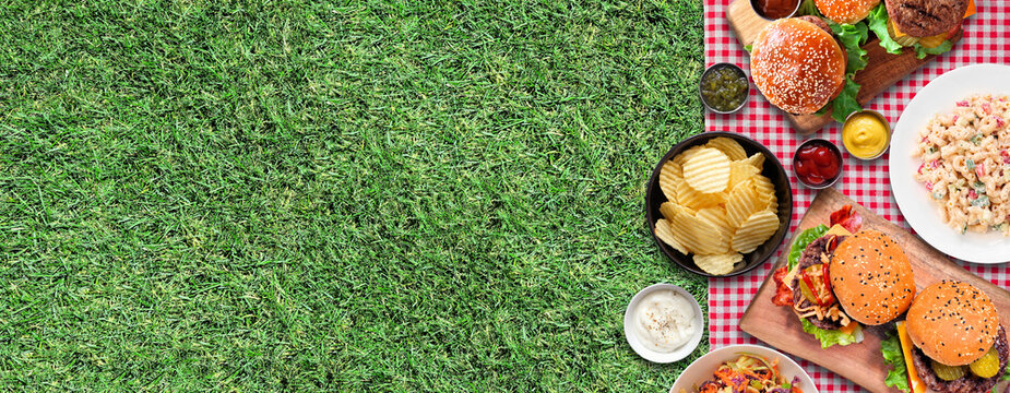 Summer outdoor BBQ side border with hamburgers, classic salads and snacks. Top view over a red and white check cloth and grass banner background. Copy space.