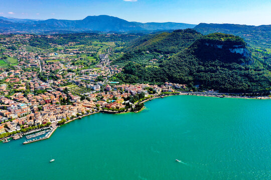 Aerial view of Lake Garda with the coastal town buildings, lush green hills, and turquoise water under a clear blue sky in Italy.