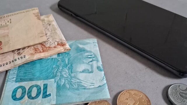Brazilian real banknotes and metal coins alongside a modern smartphone on a flat grey surface