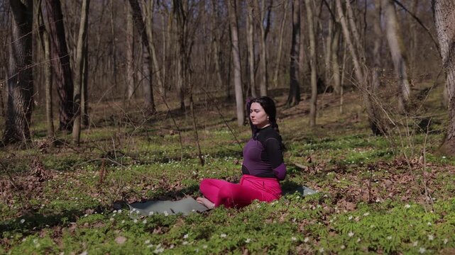 Peaceful woman in comfortable workout attire sits in yoga pose on grass stretching spine. Serene female in training clothes sits on forest lawn gently pulling arms backward to stretch upper back