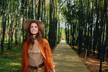 Fototapeta premium Woman in a bamboo forest on a path wearing a hat and smiling, portrait in nature. Casual travel scene walking along green trail with sunlight and relaxed joyful expression.