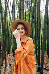Fototapeta premium Woman in bamboo forest wearing straw hat and orange shirt, smiling in portrait style. Nature travel scene with relaxed pose among tall green stalks and serene outdoor greenery.