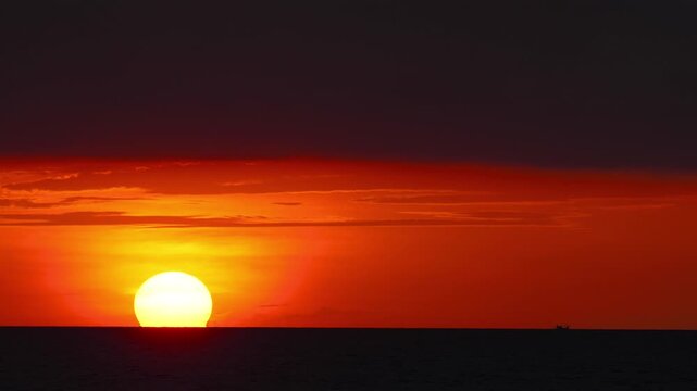 Stunning view of a massive bright sun setting over the ocean horizon. Vibrant orange and red sky with dramatic clouds. A tiny silhouette of a fishing boat is visible on the water in Phuket, Thailand.