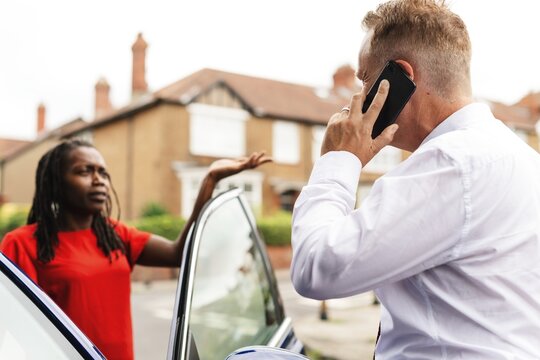 A man in a white shirt talks on a phone by a car. A woman in a red shirt stands nearby. They appear to be in a suburban neighborhood with houses in the background. Car accident concept photo.
