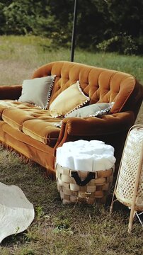 Vertical video. Elegant outdoor lounge area with vintage velvet sofa, decorative pillows and woven baskets on natural rugs during daytime wedding celebration. Cozy boho event decor in nature. Filmed