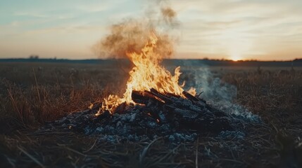Fototapeta premium A dramatic sunset illuminates a rural field where a large fire burns, casting smoke and ash into the warm twilight sky.