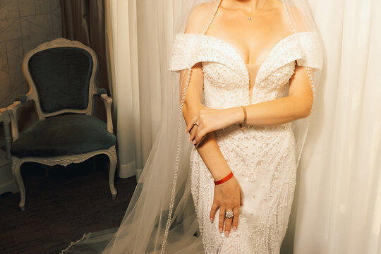 Bride in a white dress stands near bright window in a venue before wedding ceremony.
 bride stands by  window wearing  white dress with  long veil. She is preparing for wedding ceremony