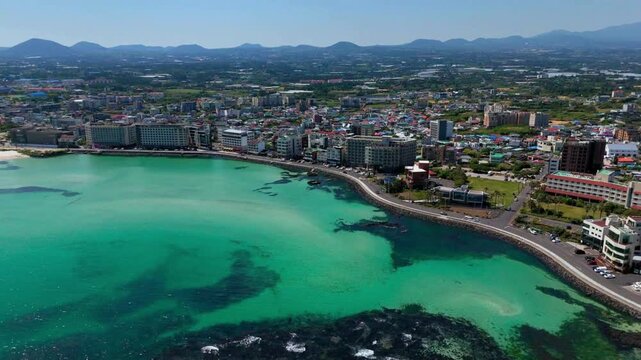 Long aerial cinematic reveal shot of Hamdeok coastal village transitioning to traditional Wondam stone fish traps with emerald sea and oreum mountains, Jeju Island, South Korea