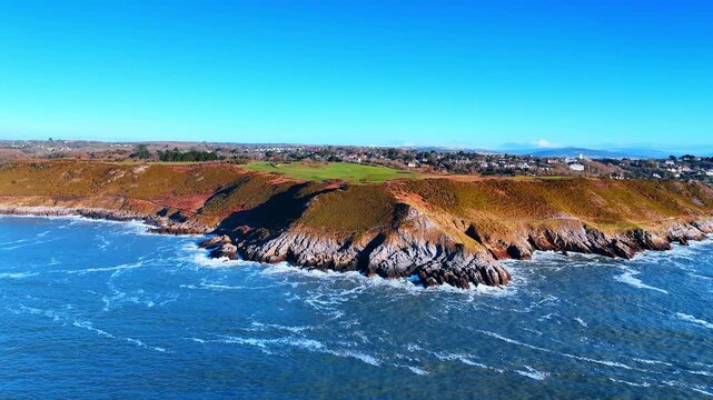Aerial View of Rocky Coastal Point with Crashing Surf and Village. White surf crashes over a jagged rocky point as a hillside village and beach huts line the shore behind it.