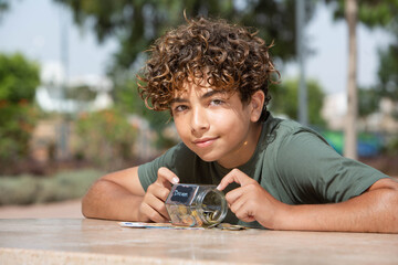 Teenager Saving Money for a Dream in Glass Jar. Close-up portrait of a smiling teenage boy with curly hair lying on a table outdoors, holding a glass jar filled with coins labeled “Dream.” The image r © NataSao