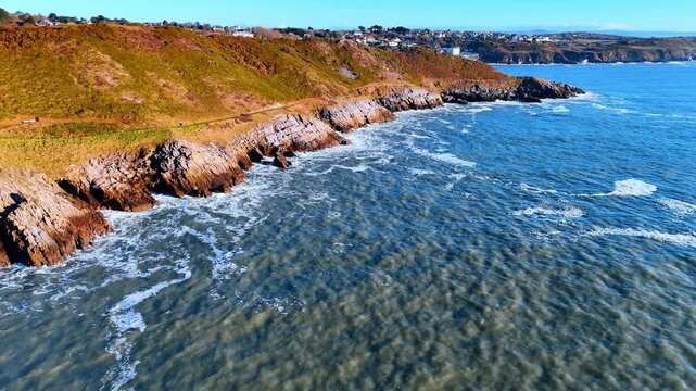 Aerial View of Welsh Coastal Cliffs with Surf and Hillside Houses. Rugged cliffs lined with a coastal path drop to a foamy sea as houses scatter across the hills along the shoreline.