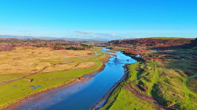 Drone View of Coastal Estuary with Green Banks and Amber Sand Dunes. A coastal estuary winds past green banks with a white building visible on the right and amber dunes on the left.