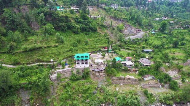 Aerial drone orbit view of a riverside villa surrounded by lush green mountains and forest landscape near beas parvati river in the scenic Manali region of Himalayas.