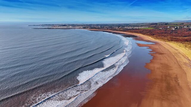 Aerial Coastal View of Sandy Shore with Parallel Breaking Waves. A broad reddish brown sandy beach curves along the coast while successive wave lines break evenly toward the shoreline.