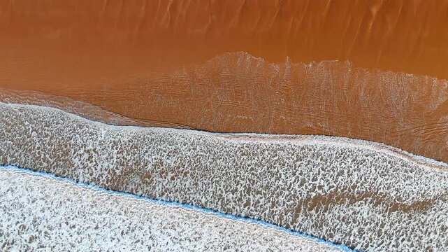 Abstract Aerial View of Ocean Waves Meeting Rust Colored Sandy Shore. A top-down aerial shows rust orange wet sand bordered by a band of white wave foam and a narrow strip of blue water.