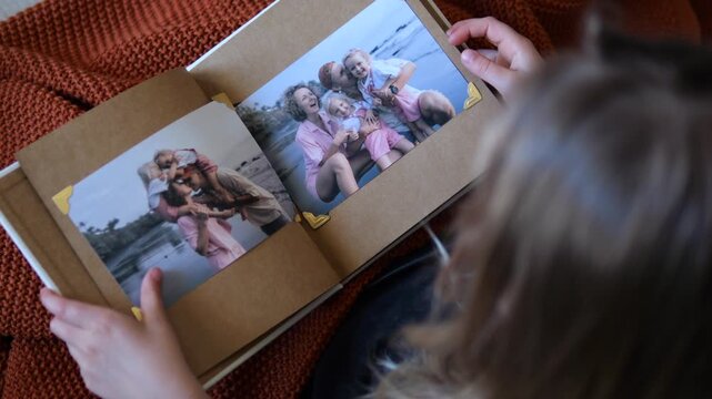 Over the shoulder view of a little girl opening a family photo album and fondly looking at old pictures, reminiscing about past happy moments and cherishing childhood memories with her relatives