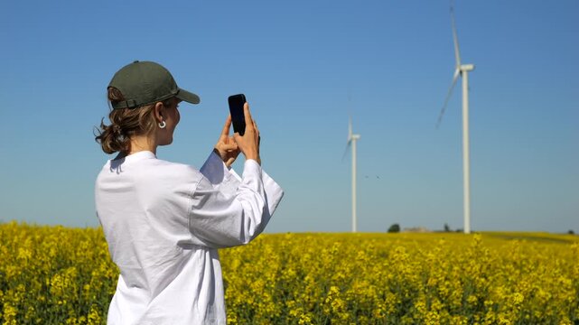 Young woman in a white shirt and cap recording with a mobile phone in a vibrant yellow rapeseed field with wind turbines generating renewable energy against a clear blue sky on a sunny day