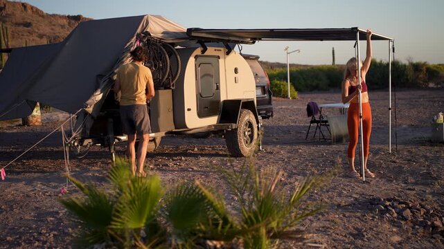 A senior man and woman open an awning on a teardrop camper trailer at sunrise on a beach campsite in Baja Mexico.