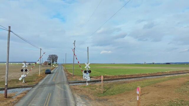 Cars wait at a train crossing where lights flash and barriers lower. The setting is a rural area with open fields in sight. The weather appears cloudy and cool.
