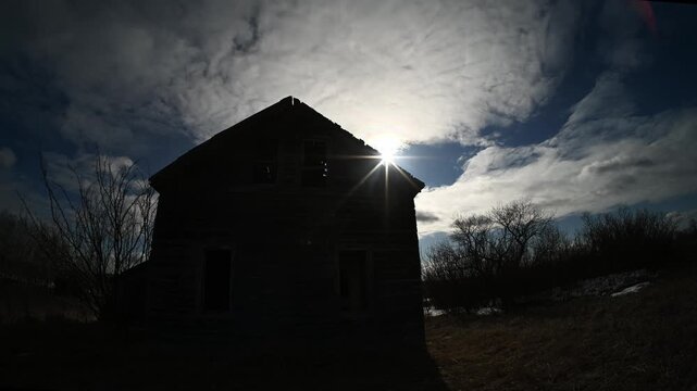Timelapse of summer clouds moving behind a dilapidated farmhouse with a dramatic sun star at the beginning of the clip
