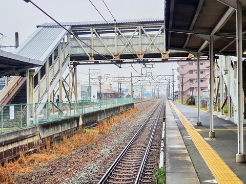 A view of a railway station platform in Japan featuring parallel tracks extending into the distance with vanishing point perspective. The scene includes a weathered steel pedestrian overpass