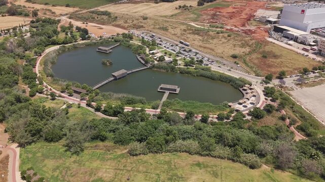 Aerial drone view of Hod Hasharon Park with scenic lake, wooden docks, green paths and nearby business complex in Israel
