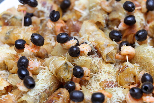 Macro close-up of savory fried pastry cigars topped with shrimp, black olives, and grated cheese