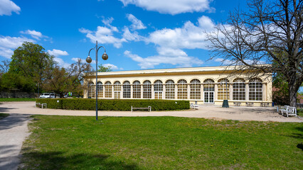 Fototapeta premium A building of the riding school is visible in Boskovice, Czechia. The structure is surrounded by grass and trees, with benches nearby. Clouds fill the blue sky above.