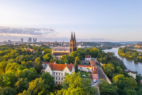 The sun rises, casting golden light over the Vysehrad Basilica of St. Peter and St. Paul in Prague. Surrounded by lush greenery, the majestic structure stands tall against the skyline.