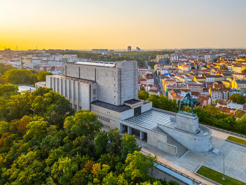The sun rises over the Vitkov National Monument where the Tomb of the Unknown Soldier is located. The cityscape of Prague is visible in the background with buildings and trees.