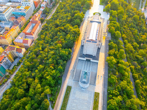 The National Monument at Vitkov stands tall during sunrise. The Tomb of the Unknown Soldier can be seen surrounded by green trees and city buildings, marking an important historical site in Prague.