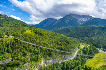 Fototapeta premium Highline 179 is a pedestrian suspension bridge in Austria. It connects a castle's ruins with a Roman fortress. Visitors walk across the bridge over a wide river gorge surrounded by mountains.