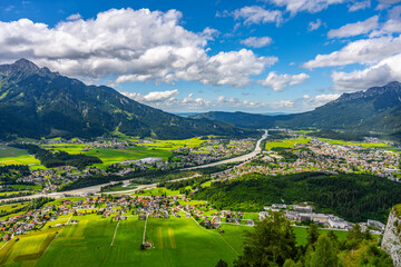 Fototapeta premium A wide view shows Reutte in Tyrol, Austria. Green fields and houses spread out below. Mountains rise in the background under a blue sky with clouds. The river flows through the valley.