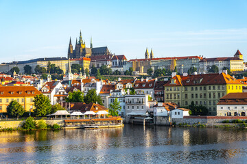 Obraz premium Prague Castle stands tall above the city, with the Vltava River flowing below. Buildings line the riverbank, showing a mix of architecture against a clear sky during the day.