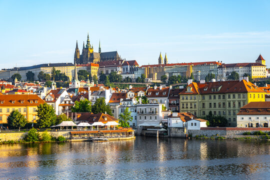 Prague Castle stands tall above the city, with the Vltava River flowing below. Buildings line the riverbank, showing a mix of architecture against a clear sky during the day.