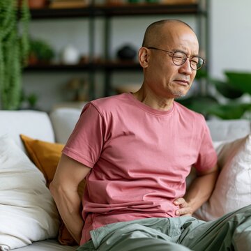 Bald man wincing while holding lower back on living room sofa