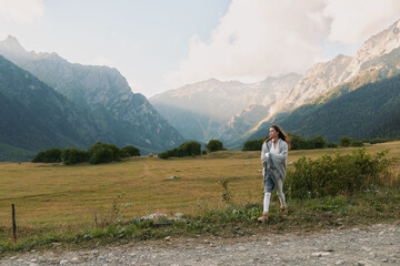 Fototapeta premium Young woman walks along a gravel road through a mountain valley, wearing a denim jacket and scarf, enjoying the wide landscape and fresh air as distant peaks loom