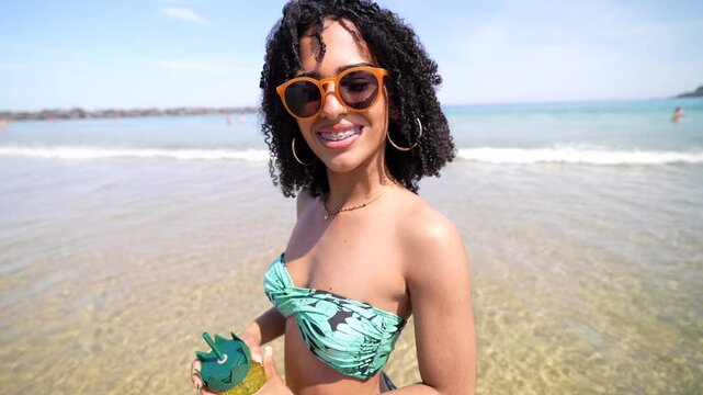 Beautiful young african american woman with curly hair and sunglasses smiling and dancing in the sea, holding a pineapple shaped cup during her summer vacation on a sunny day at the beach