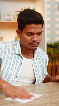 Indian man on home table wipes desk with napkin makes space and opens notebook for neat work routine. Arabian boy on home table brings order then writes notes with calm focus during indoor planning.