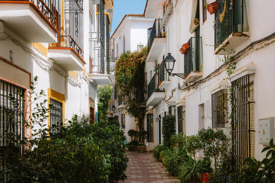 Narrow street with traditional white buildings featuring wrought iron balconies and green plants, creating a picturesque ambiance in Marbella, Andalucia, Spain