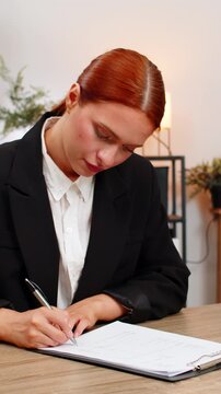 Young businesswoman at home office turns pages of contract reading each line and signing with hand. Freelancer girl at table finalizes paperwork sealing deal satisfied with legal clarity project start
