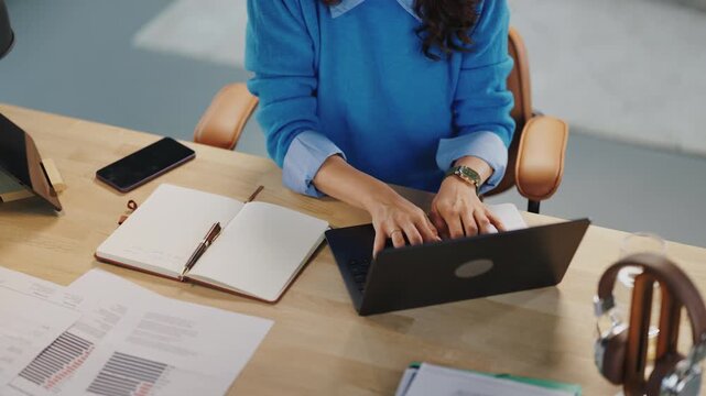 A person is typing on a laptop at a wooden desk. There is a notebook, a pen, and a phone on the desk. Papers are spread out, showing data and graphs.