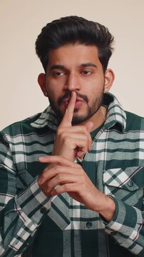 Shh be quiet please. Young Indian man presses index finger to lips makes silence gesture sign do not tells secret, stop gossip. Handsome bearded Hindu boy isolated alone on beige studio background
