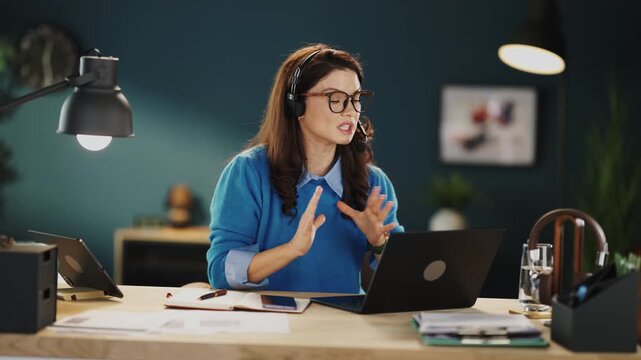 A person is sitting at a wooden desk using a laptop. She wears a headset and smiles while speaking. Papers and a glass of water are on the desk. The setting is bright and modern.