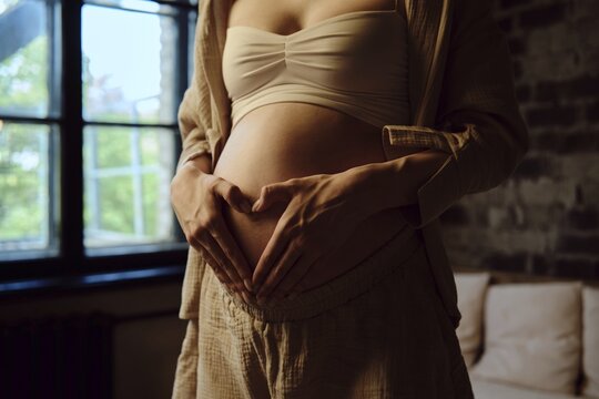 In a cozy home setting, a pregnant woman stands near a window, gently placing her hands in a heart shape on her rounded belly. Soft light illuminates the room, highlighting her joyful anticipation