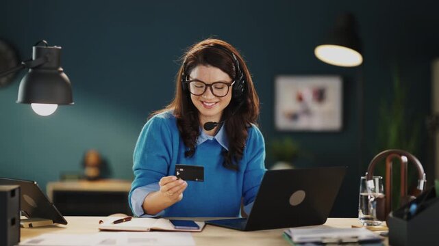A woman sits at a desk using a laptop while holding a card. She is focused on her work. A lamp and a drink are on the desk. The room has a blue wall.