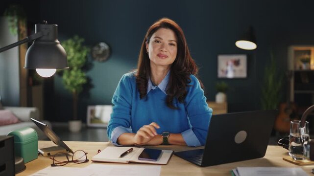 A woman writes in a notebook while sitting at a desk with a laptop and plant around her. She appears focused as she takes notes and uses a pen to jot down ideas.