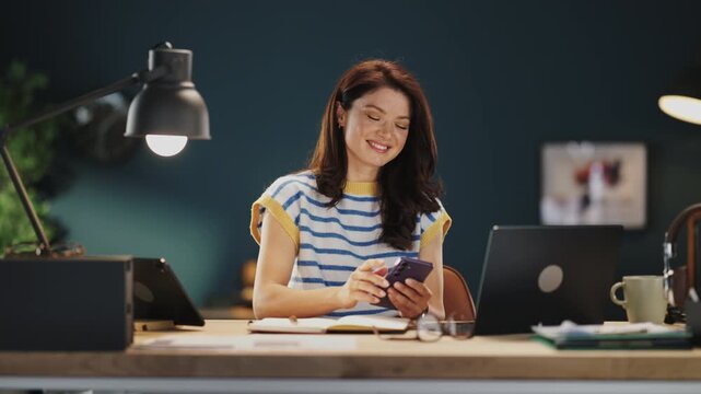 A woman sits at a desk in a home office. She smiles while looking at her phone. A laptop is nearby. The room is well lit and has simple decor with a green background.