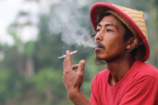 Asian Indonesian rural man in red shirt and traditional conical straw hat smoking a cigarette outdoors.