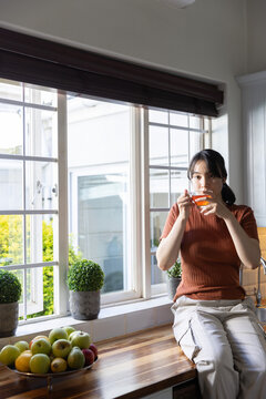 Asian woman holding teacup sipping tea on kitchen counter by window with apple bowl, copy space